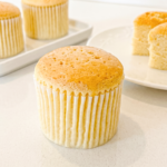 A honey castella muffin in a white liner sits on a light counter, with two more muffins and two cut pieces of a muffin in the background.