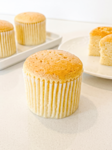 A honey castella muffin in a white liner sits on a light counter, with two more muffins and two cut pieces of a muffin in the background.