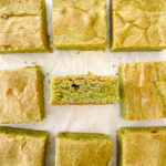 Several square-cut matcha blondies with golden tops and green interiors are arranged on white parchment paper. One blondie is on its side, showing its texture.