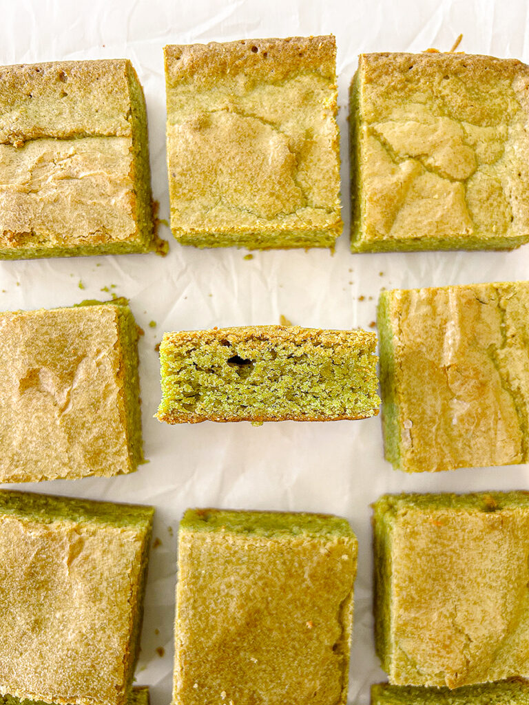 Several square-cut matcha blondies with golden tops and green interiors are arranged on white parchment paper. One blondie is on its side, showing its texture.