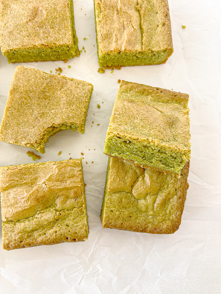 Six square matcha blondies with a light brown top and green centre, scattered on white parchment paper, with one piece having a bite taken out.