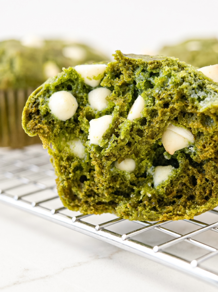 A green matcha muffin, split open to show its crumb and white chocolate chips, sits on a metal cooling rack.