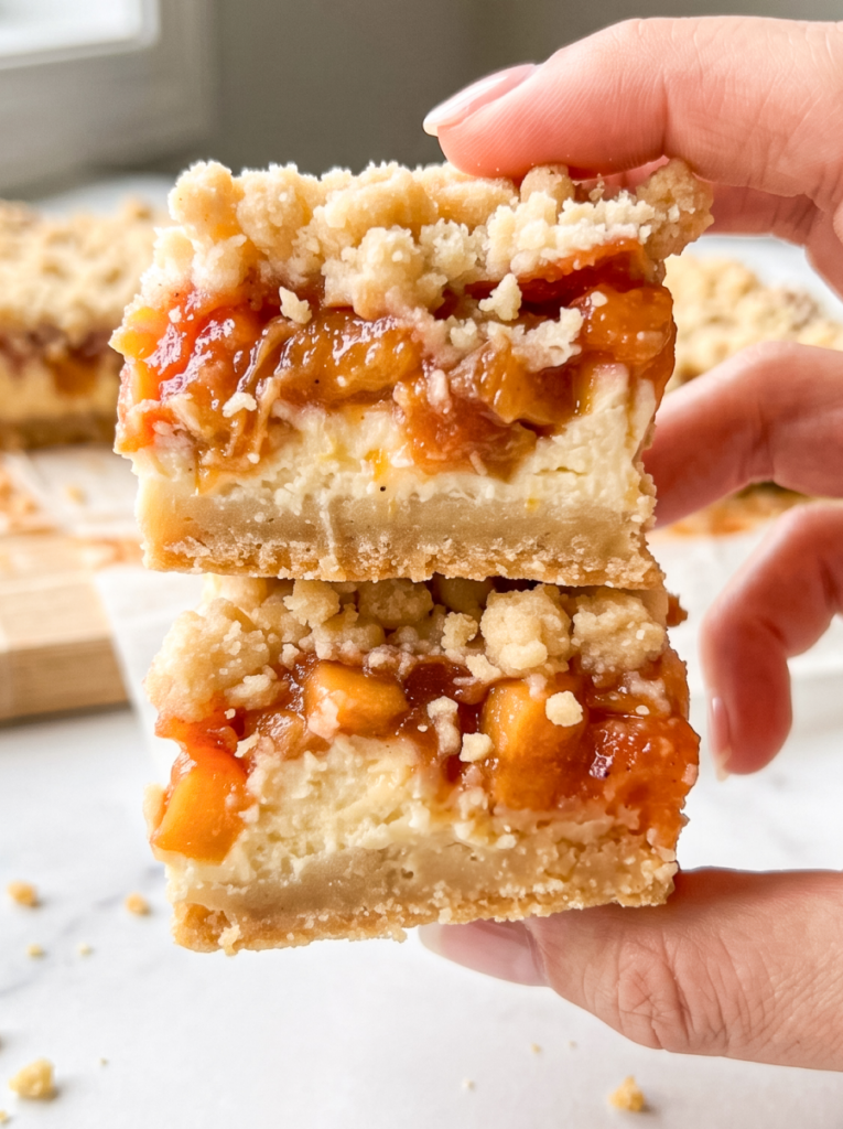 A hand holds two stacked peach cheesecake bars, showing their shortbread crust, creamy cheesecake, peach topping, and crumble.