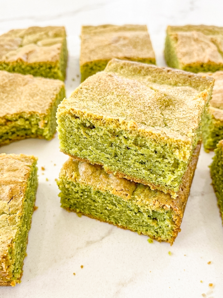 Green matcha blondies with golden-brown tops are arranged on a white surface, with two squares stacked in the foreground.