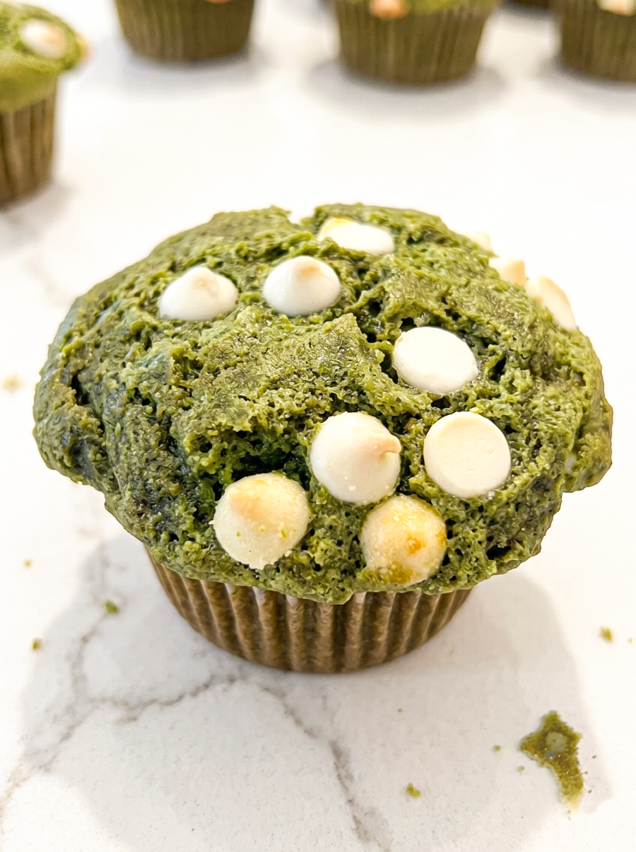 A close-up of a green matcha muffin with white chocolate chips on top, sitting in a brown paper liner on a white marble surface.