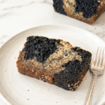 A slice of marbled black sesame banana bread with dark grey and light brown colours is on a white plate with a fork beside it.