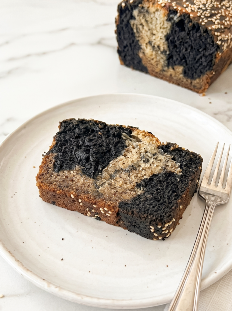 A slice of marbled black sesame banana bread with dark grey and light brown colours is on a white plate with a fork beside it.