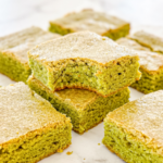 Square matcha blondies with a golden top crust are arranged on a marble surface; one stacked blondie has a bite taken out.
