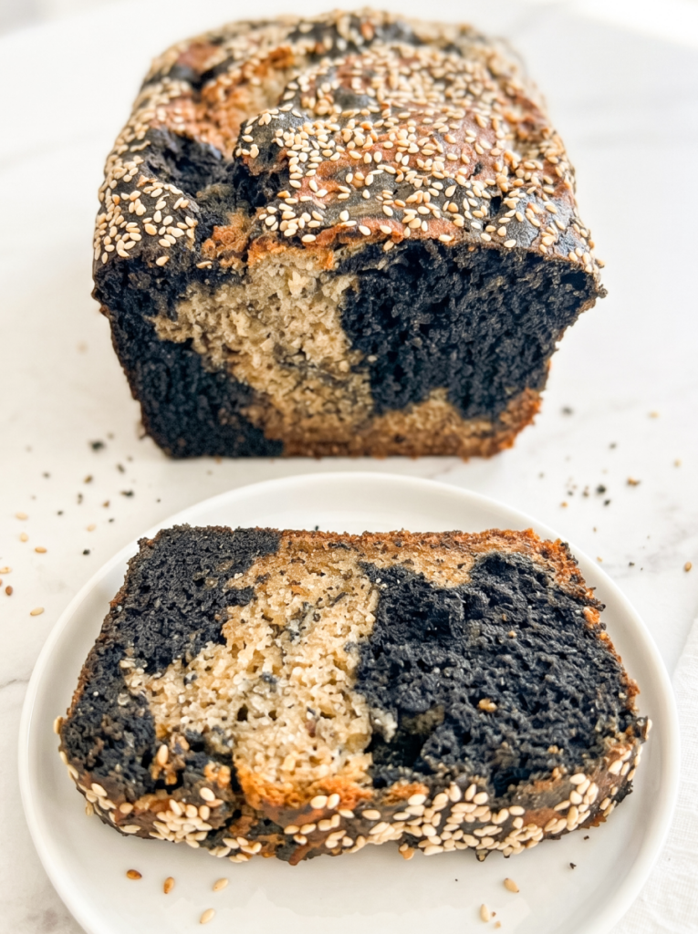 A marbled loaf of black sesame banana bread and a slice on a white plate are topped with white sesame seeds.
