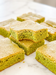 Square matcha blondies with a golden top crust are arranged on a marble surface; one stacked blondie has a bite taken out.