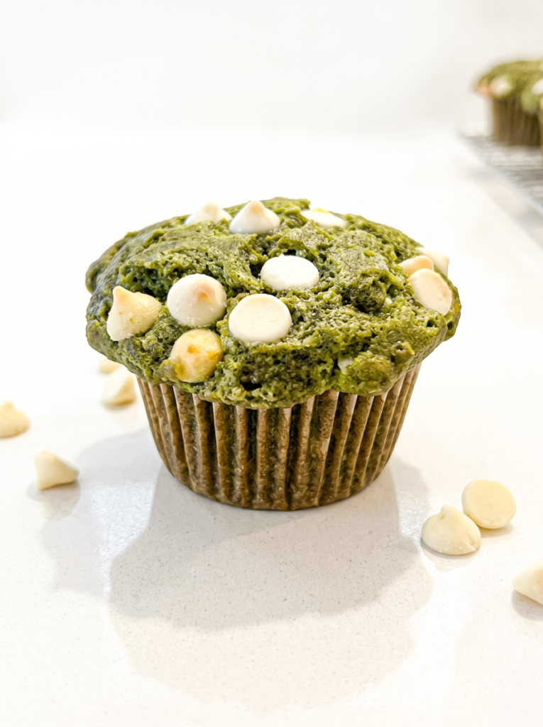 A green matcha muffin topped with white chocolate chips sits on a white surface, with scattered chips nearby and more muffins out of focus.