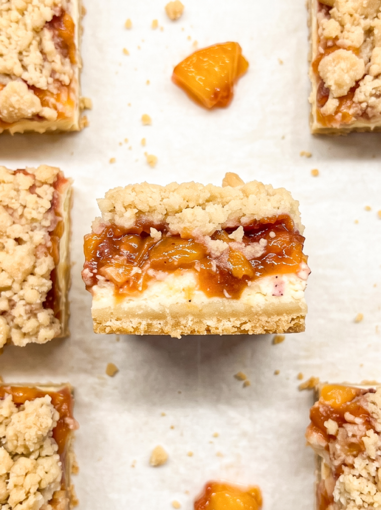 I see a close-up of a peach cheesecake bar with a crust, cream cheese, peach topping, and streusel, with other bars visible.