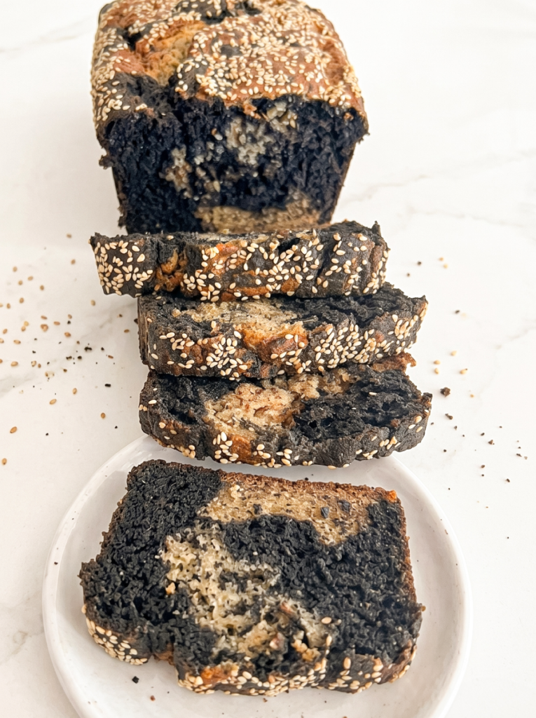 A sliced loaf of black sesame banana bread sits on a white marble surface, showing its dark and light marbled interior with white sesame seeds on top.