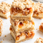 Three peach cheesecake bars are stacked on a white marble counter, showing their crumb topping, peach layer, and cheesecake.