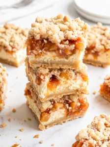 Three peach cheesecake bars are stacked on a white marble counter, showing their crumb topping, peach layer, and cheesecake.