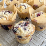 Bakery style blueberry muffins with streusel topping sit on a wire rack, with loose blueberries on the marble counter.