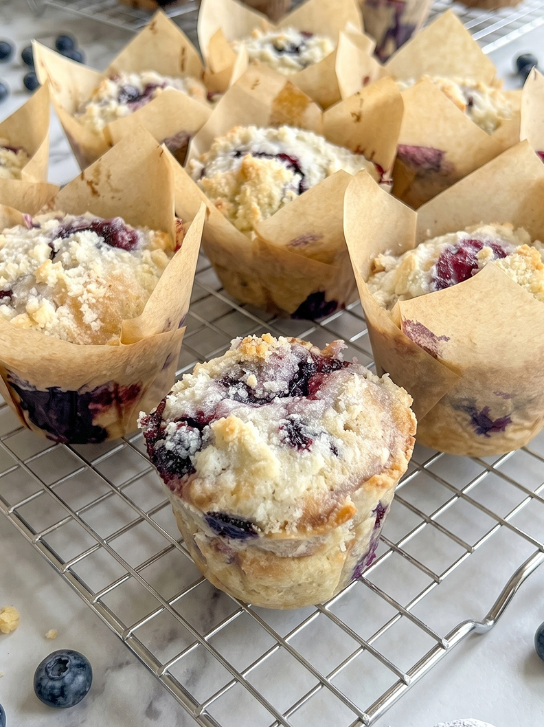 Bakery style blueberry muffins with streusel topping sit on a wire rack, with loose blueberries on the marble counter.