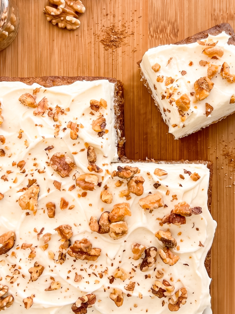 An overhead view of a frosted carrot sheet cake topped with walnuts on a wooden board, with one slice removed.