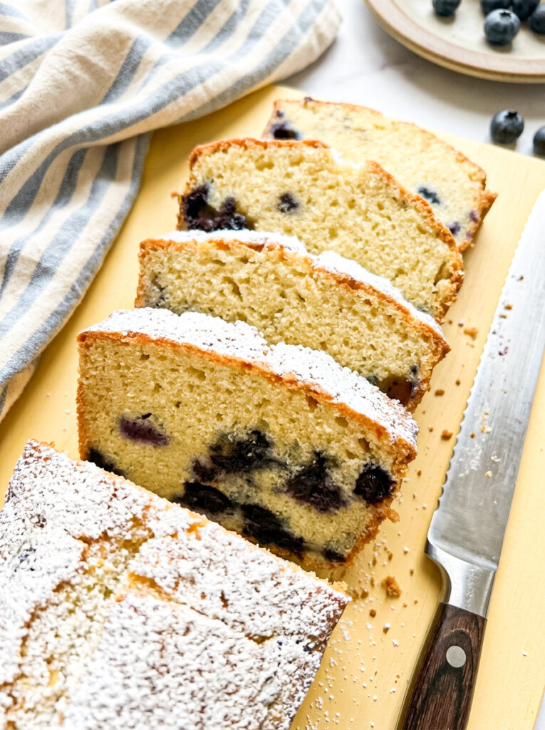 Slices of blueberry pound cake dusted with powdered sugar sit on a wooden board beside a knife.