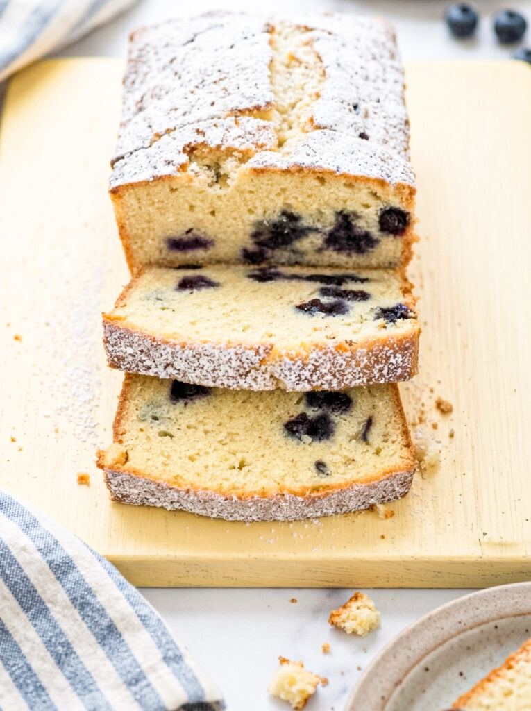 A loaf of blueberry pound cake dusted with powdered sugar, cut into slices on a wooden board.
