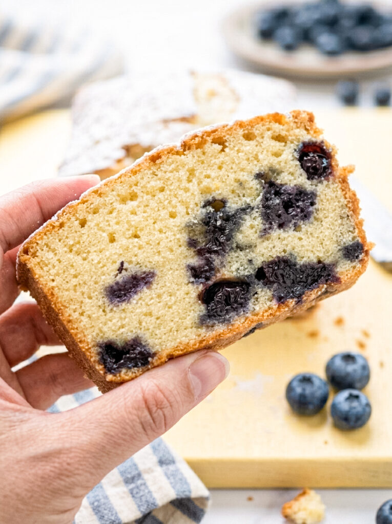 A hand holds a slice of blueberry pound cake filled with cooked blueberries over a yellow cutting board.