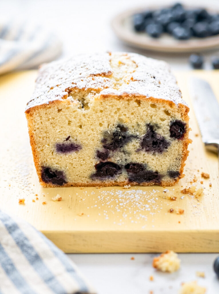 A sliced blueberry pound cake dusted with powdered sugar sits on a wooden cutting board with berries in the background.