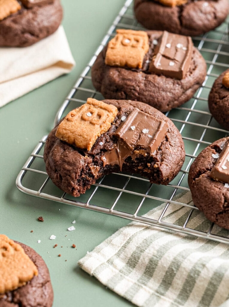 Chocolate cookies topped with Biscoff pieces and chocolate squares on a wire rack, with one cookie showing a melted chocolate centre.