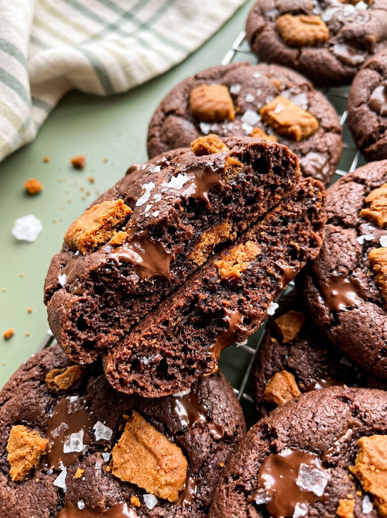 Thick chocolate cookies topped with Biscoff pieces, melted chocolate, and sea salt, with one broken cookie showing a soft chocolate centre.