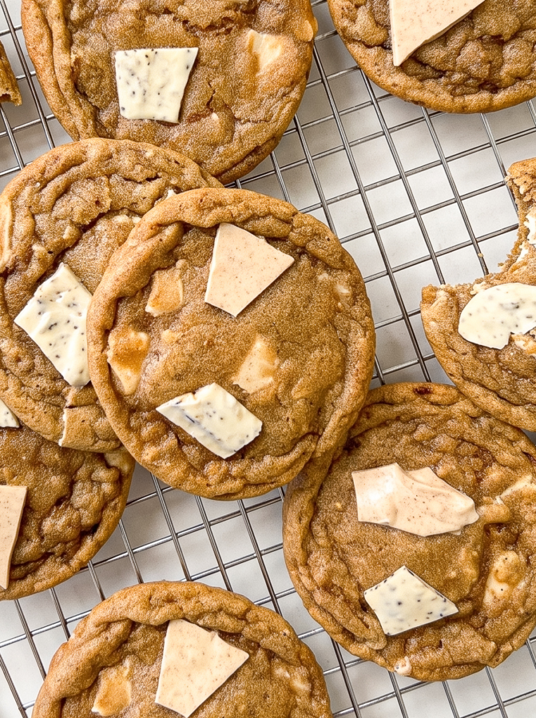 Several round, light brown pumpkin spice latte cookies with white and tan chocolate pieces are arranged on a metal cooling rack.