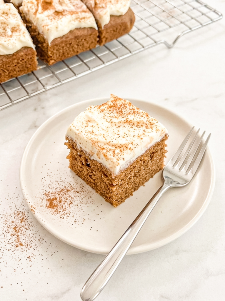 A square slice of chai spiced sheet cake with white frosting and spice sprinkles sits on a plate with a fork, with more cake in the background.