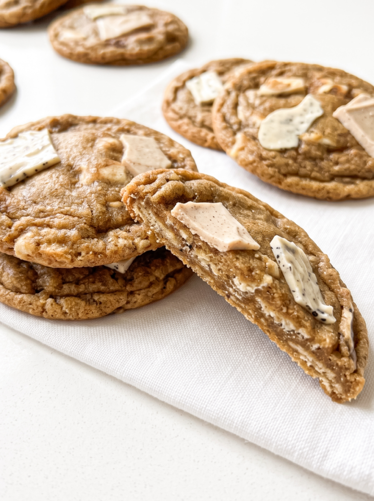 Several chewy pumpkin spice latte cookies with white and light brown chocolate pieces are arranged on a white cloth, with one cookie broken open.