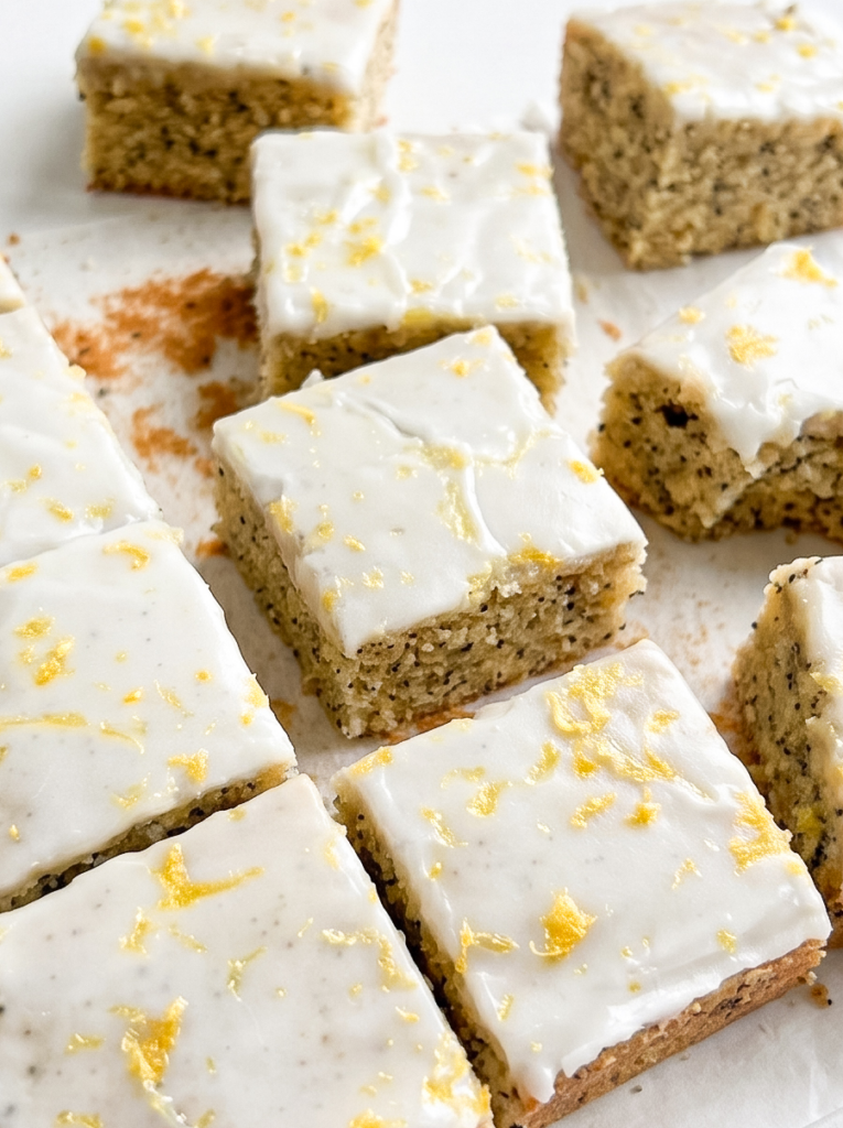 Square lemon poppyseed blondies with white glaze and yellow lemon zest are arranged on a white surface.