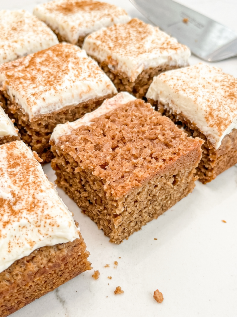 Square slices of chai spiced sheet cake with white frosting dusted with spice are arranged on a white surface, with one slice showing the cake texture.