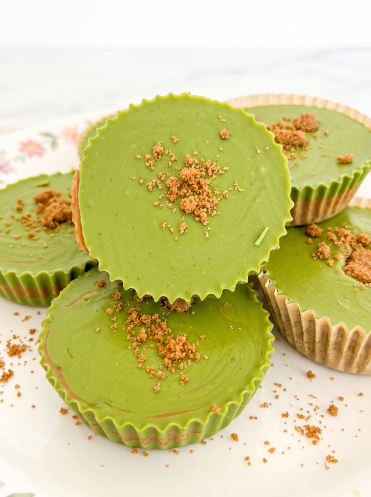 A close-up of several round, green matcha biscoff cups in fluted liners, topped with brown crumbs, stacked on a white floral plate.