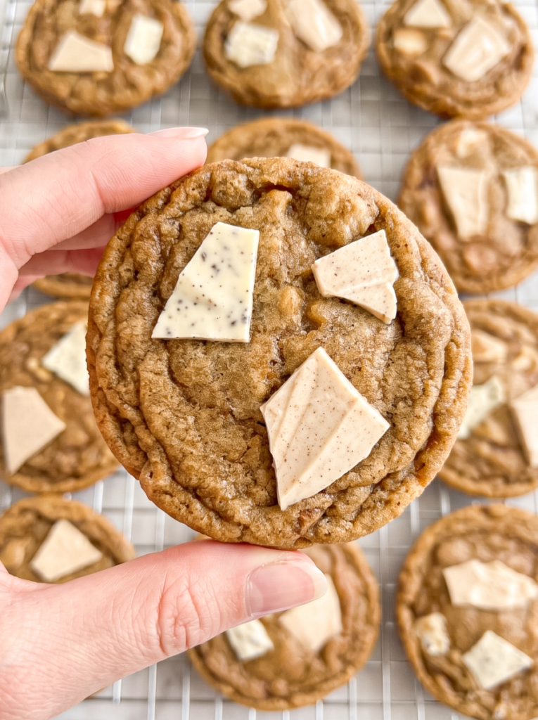 A hand holds a chewy pumpkin spice latte cookie topped with white and light brown chocolate chunks, with more cookies on a cooling rack.