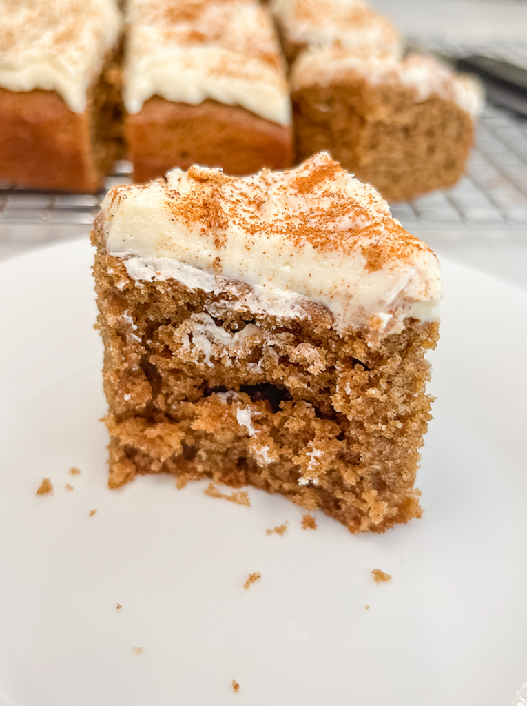 A square slice of chai spiced sheet cake with white frosting and a dusting of spice, with a bite taken from the front.