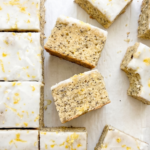 Square lemon poppyseed blondies with white lemon glaze and bright yellow lemon zest are arranged on white parchment paper.