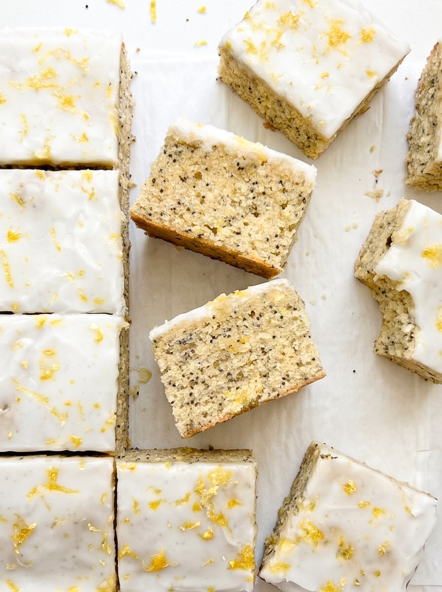 Square lemon poppyseed blondies with white lemon glaze and bright yellow lemon zest are arranged on white parchment paper.