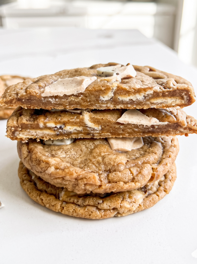 A stack of three golden brown pumpkin spice latte cookies with white and pumpkin spice coloured chunks, the top cookie broken to show its chewy centre.
