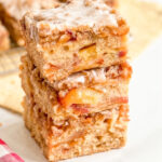 Sliced peach cobbler blondies with a streusel topping and white glaze sit on a yellow cloth next to fresh peach slices.