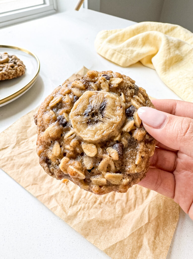 Banana walnut chocolate oatmeal cookie topped with a baked banana slice, held in hand.